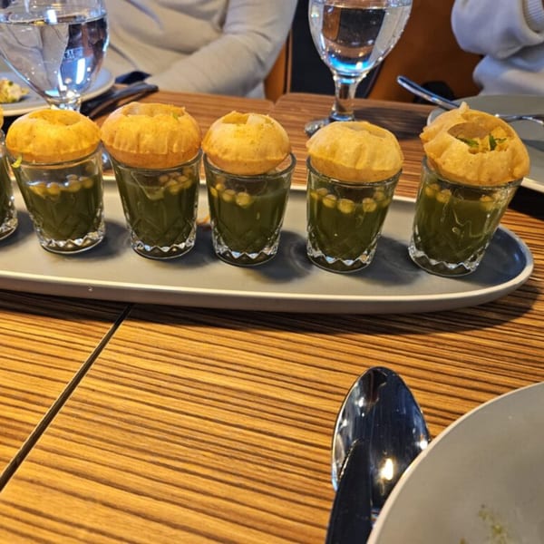 Six shot glasses of Mausam Indian Flavors pani puri, filled with green liquid and chickpeas.