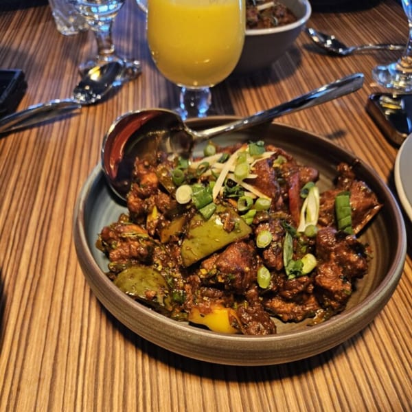 A bowl of Mausam Indian Flavors' chili chicken, garnished with green onions and ginger, on a wooden table.